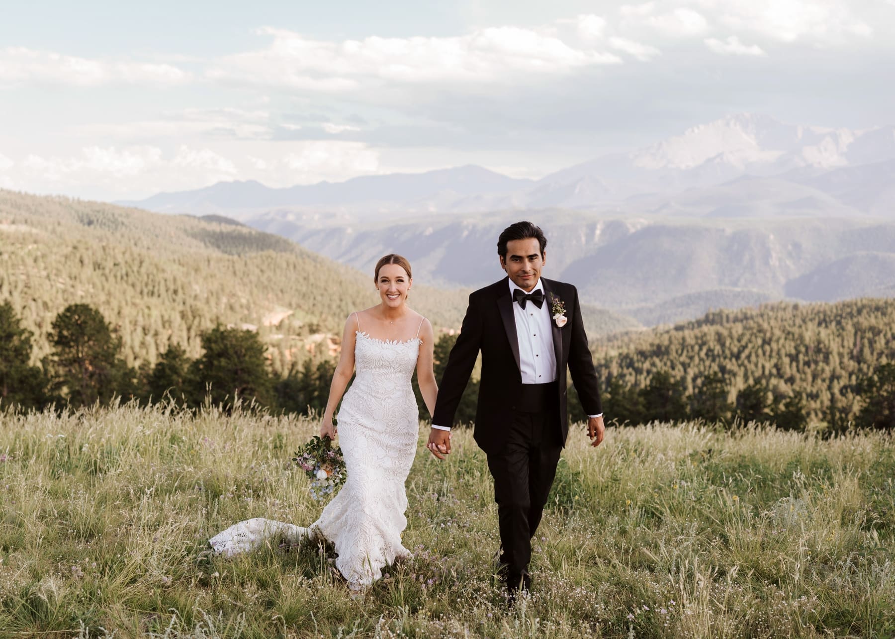Bride and groom holding hands and walking in field in front of Colorado mountains 