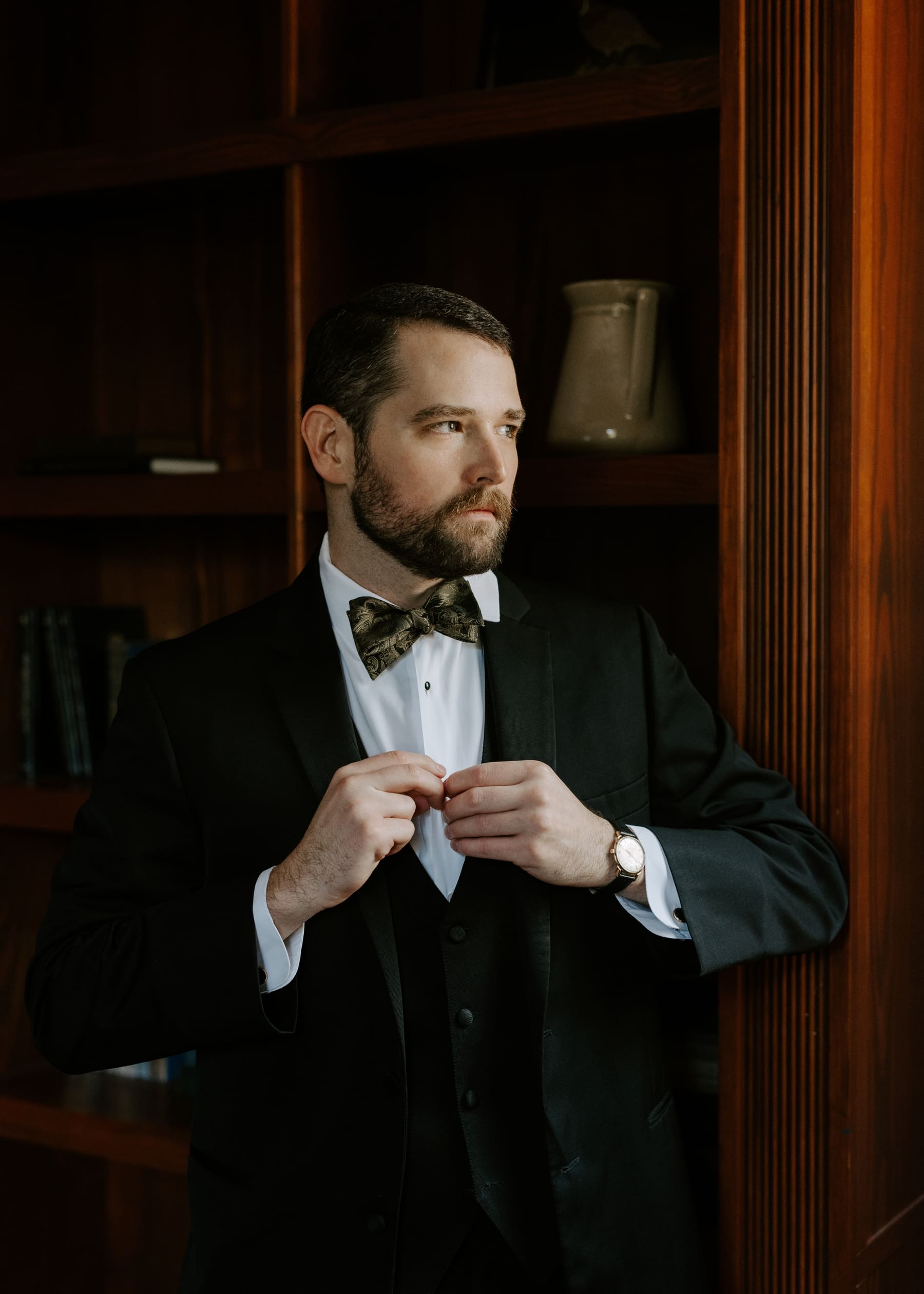 Groom buttoning shirt of tuxedo and looking out window