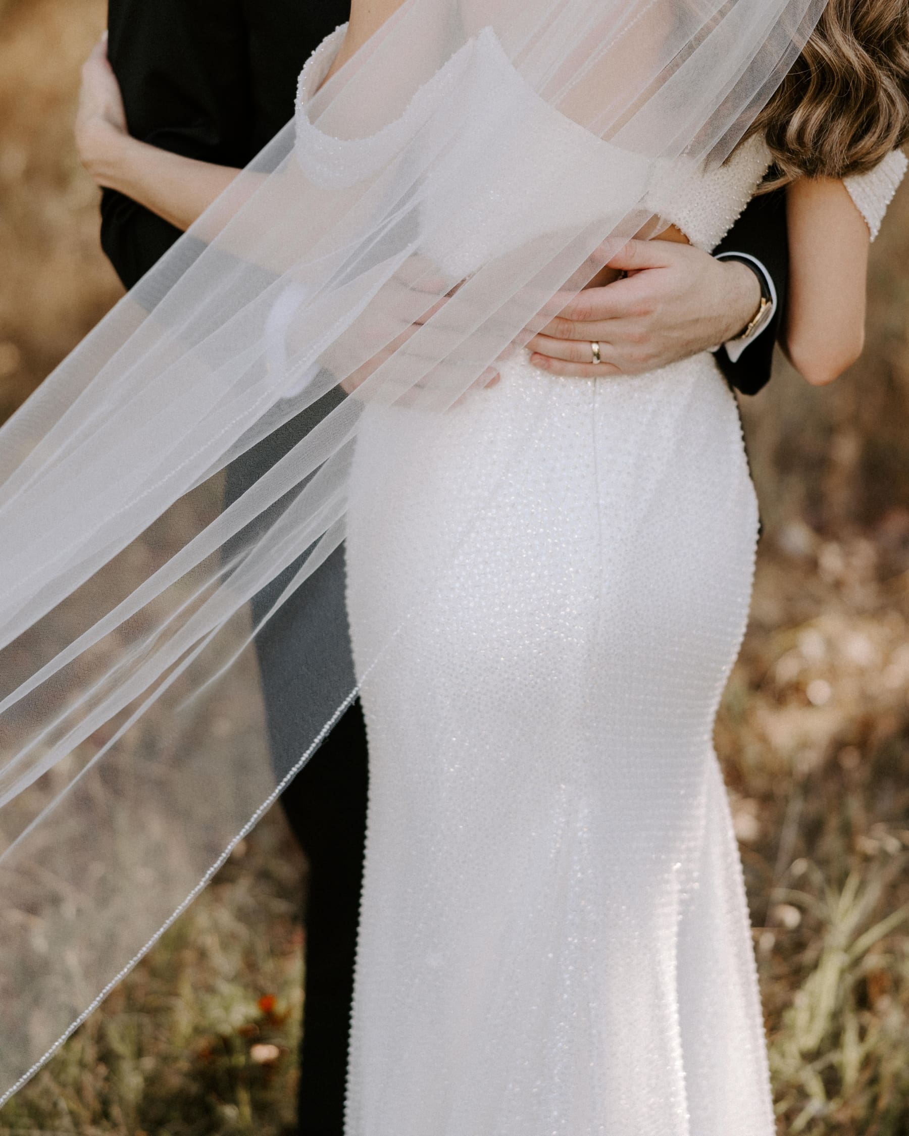 Groom's hands on bride's lower back with veil flowing in the wind