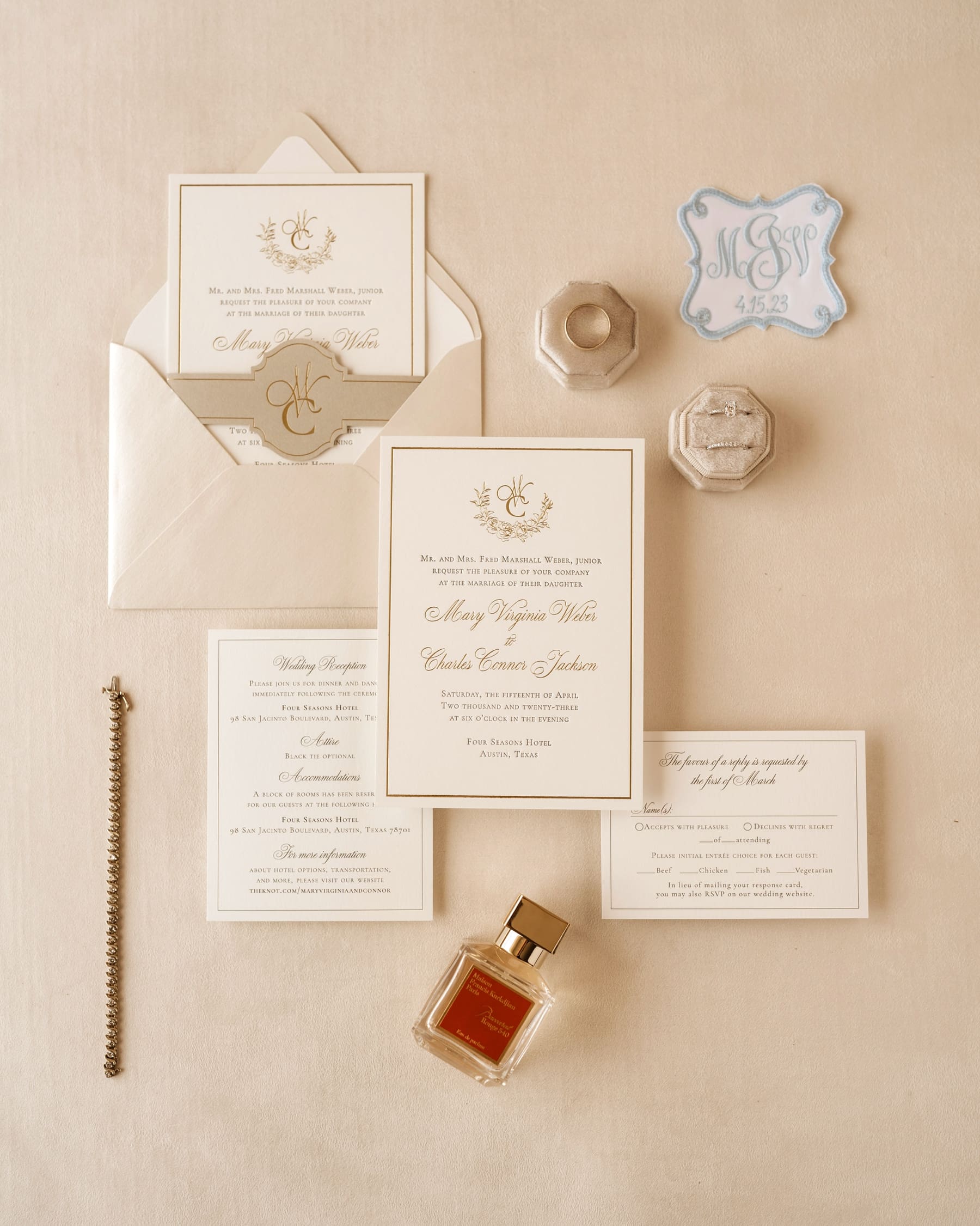 Flat lay featuring formal invitation suite with wedding rings, bracelet, perfume, and monogram patch 