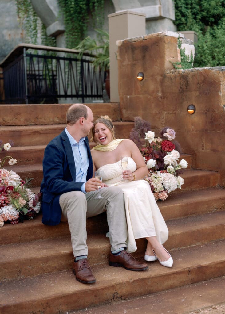 Couple sitting on rustic stone steps at Villa Antonia, laughing together with champagne in hand and floral arrangements around them.