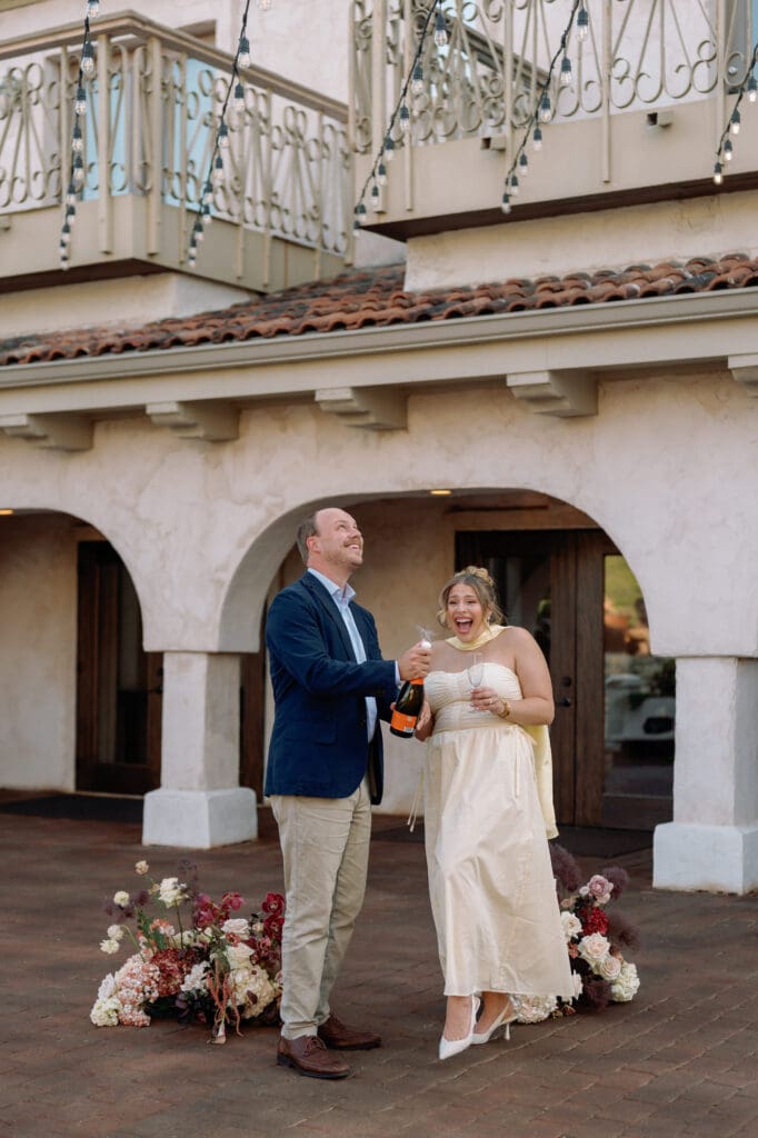 Couple celebrating their first anniversary at Villa Antonia, laughing as they pop a bottle of champagne under string lights surrounded by romantic florals.