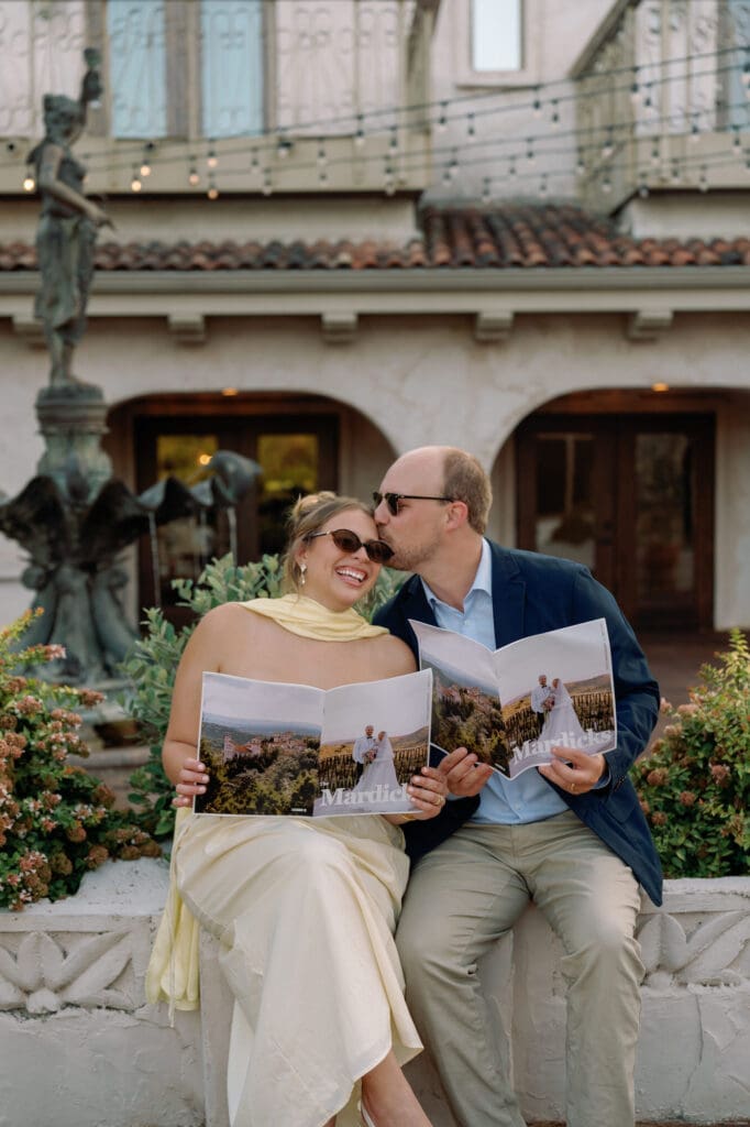 Couple sitting together, smiling and holding custom magazines recapping their first year of marriage, with the groom kissing the bride’s forehead.